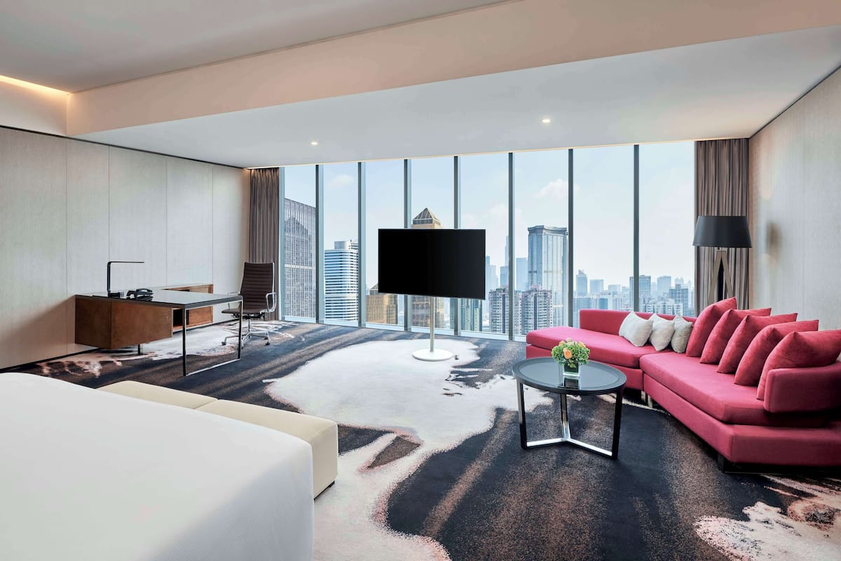 A modern hotel room with a large window, a flat-screen TV, a red sofa, and a desk with a chair.