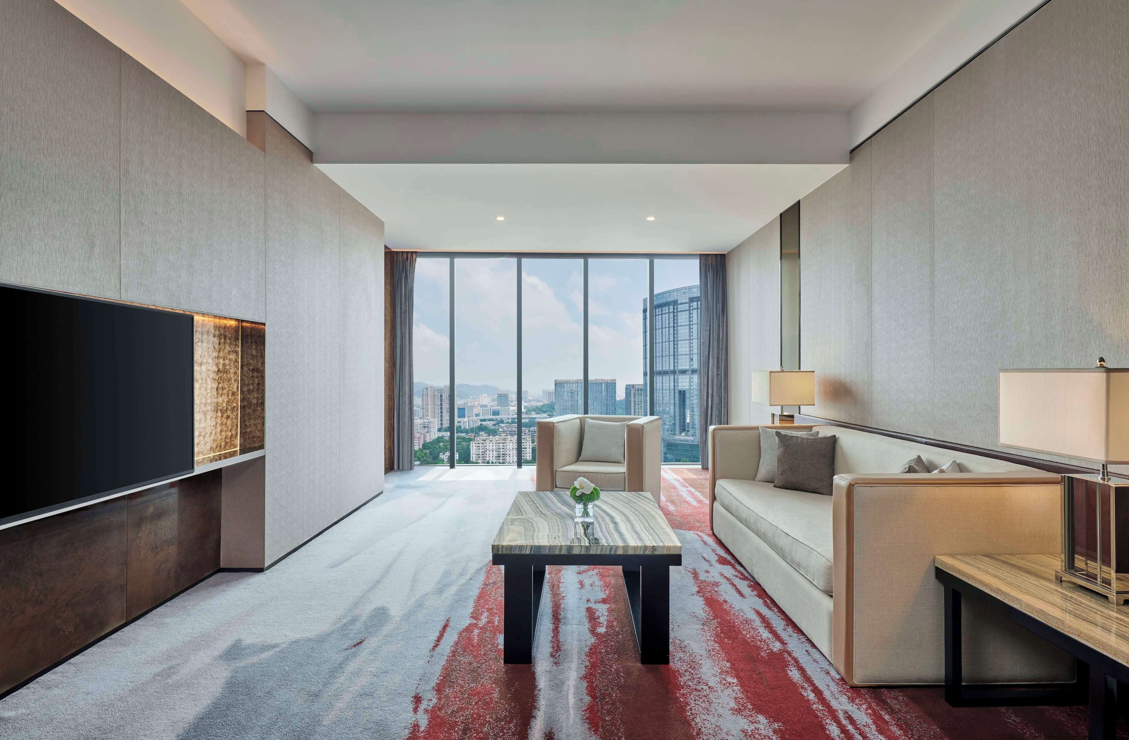 A modern hotel room with a large TV, a sofa, a coffee table, and a view of the city skyline.