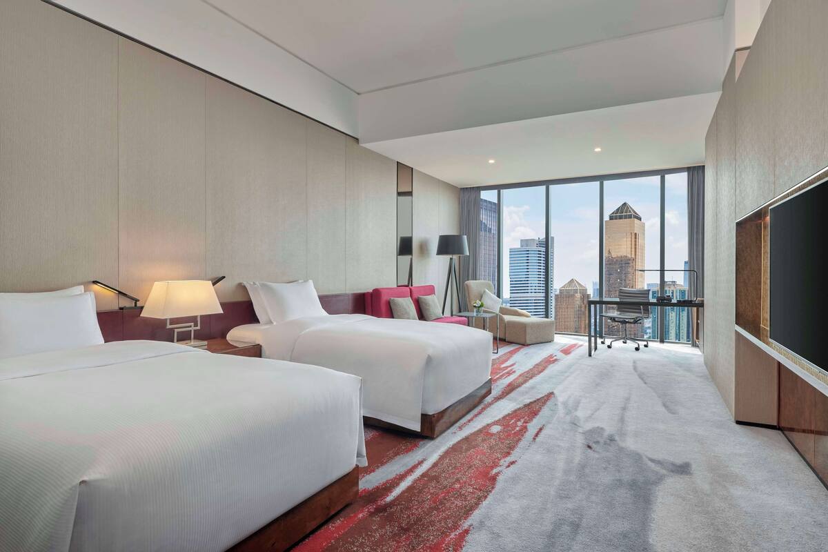 A hotel room with two beds, a desk, a chair, and a large window offering a city view.