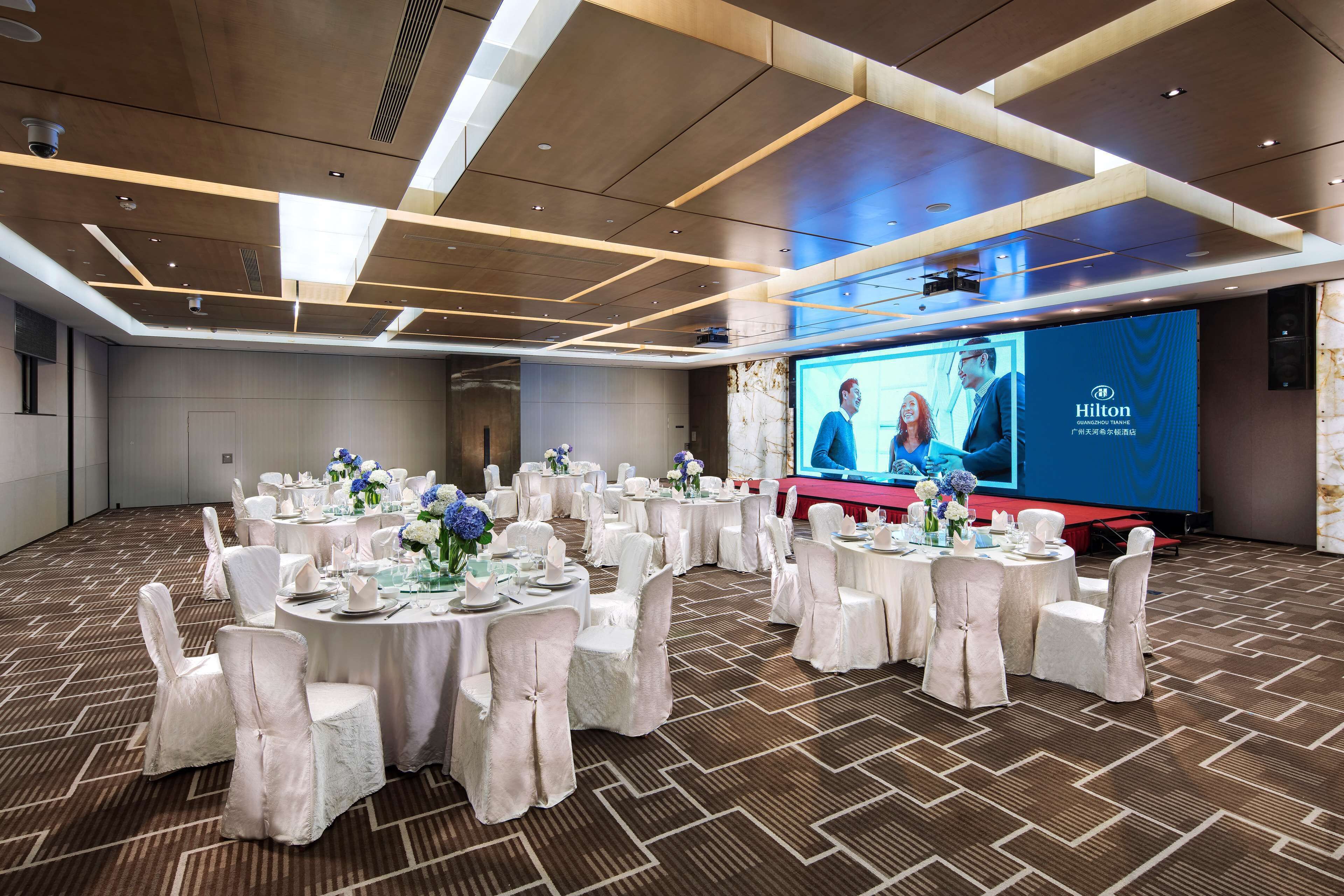 A conference room with round tables, white chairs, and a large screen displaying a Hilton logo.