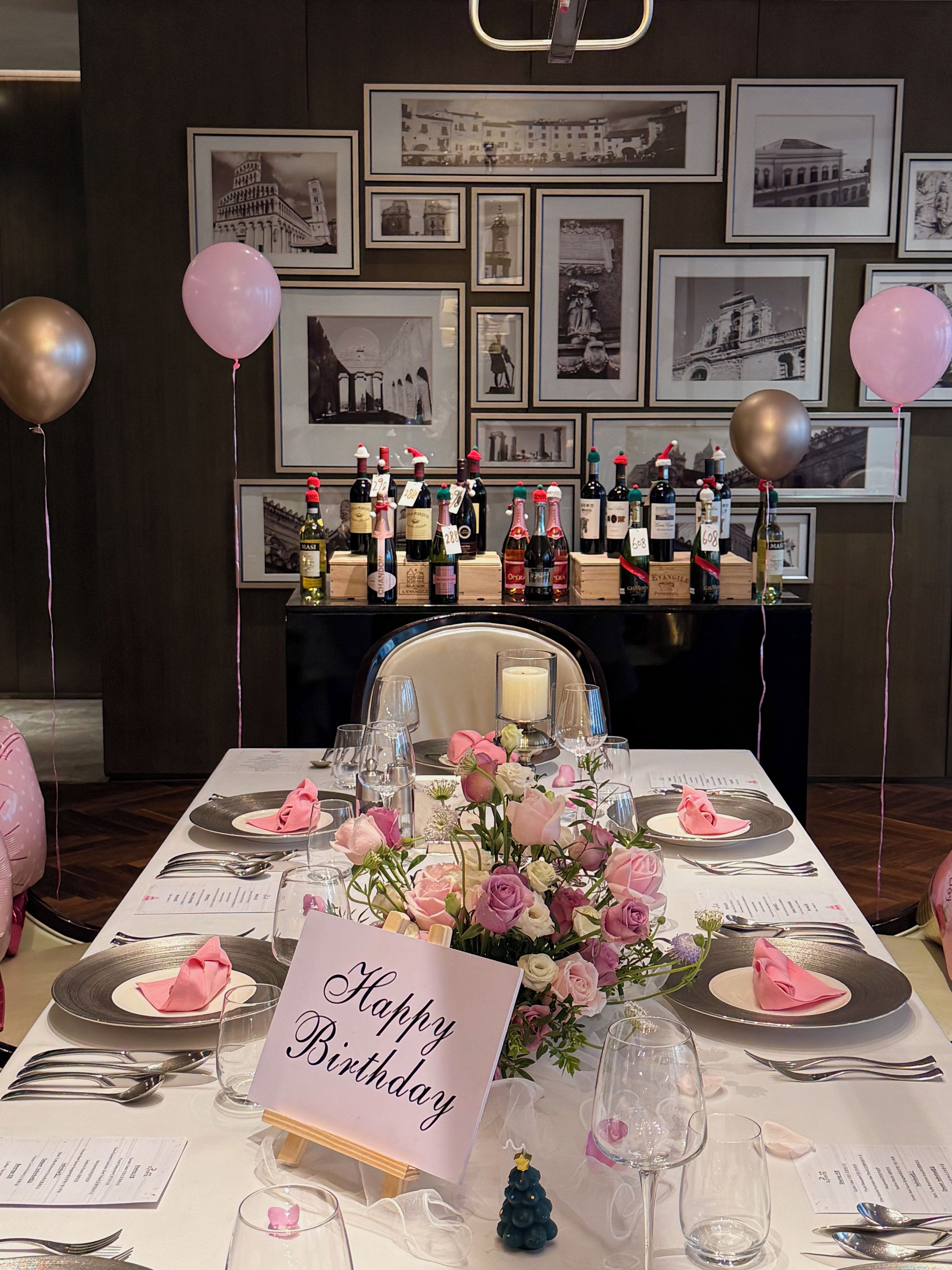 Een eettafel gedekt voor een verjaardagsviering, met een 'Happy Birthday' bord, roze ballonnen en een aantal flessen wijn.