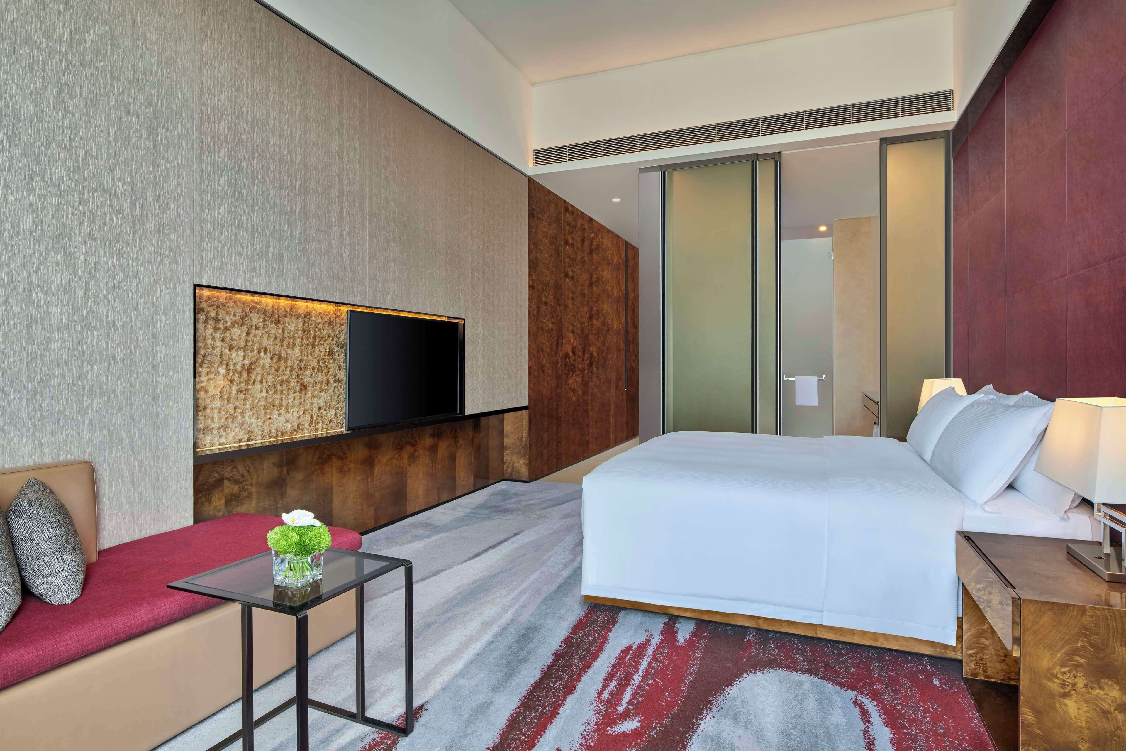 Una habitación de hotel moderna con una cama grande, un televisor de pantalla plana montado en la pared, una mesita con un jarrón y un sofá rojo.