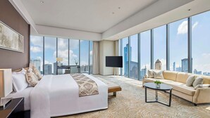 Una habitación de hotel moderna con una cama grande, un sofá, una mesa de centro y un televisor. La habitación tiene ventanas amplias con vistas a la ciudad.