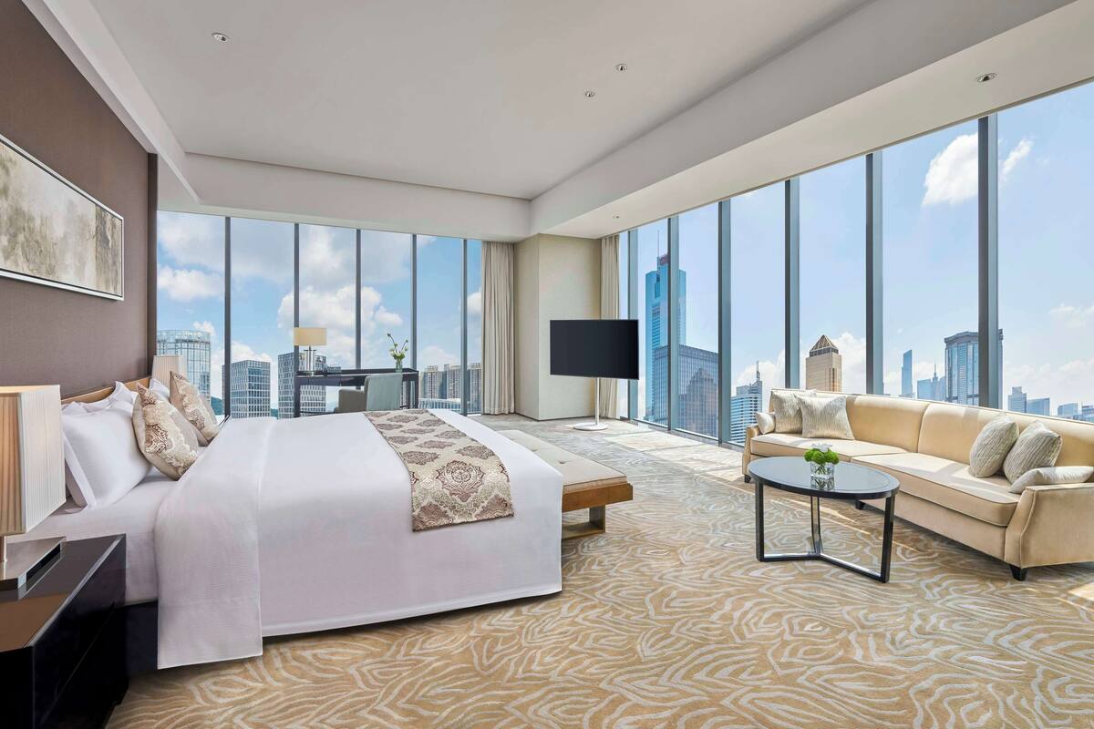 A modern hotel room with a large bed, a sofa, a coffee table, and a TV. The room has large windows with a city view.