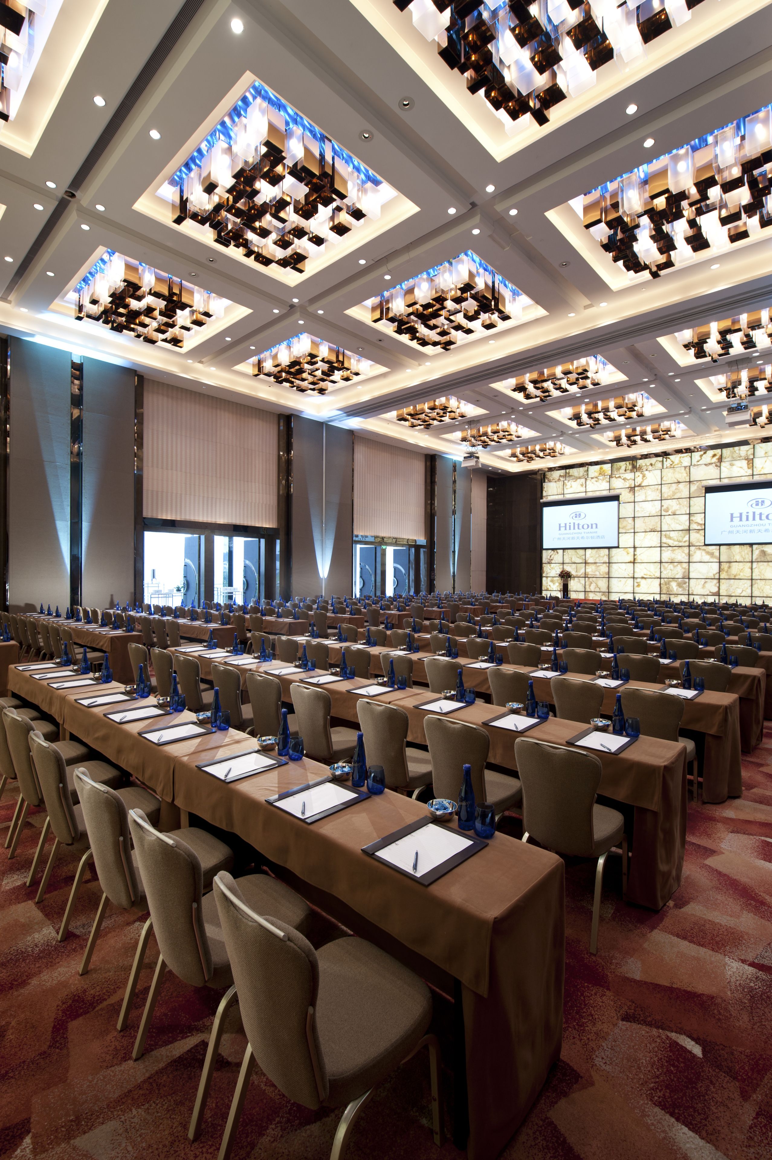 Een vergaderzaal met rijen tafels, stoelen en een groot scherm waarop het Hilton-logo wordt getoond.