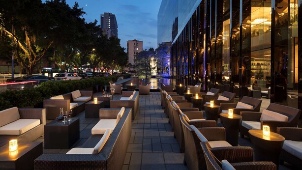 Un espacio al aire libre con mobiliario de mimbre y velas, con vista a una calle de la ciudad al atardecer.