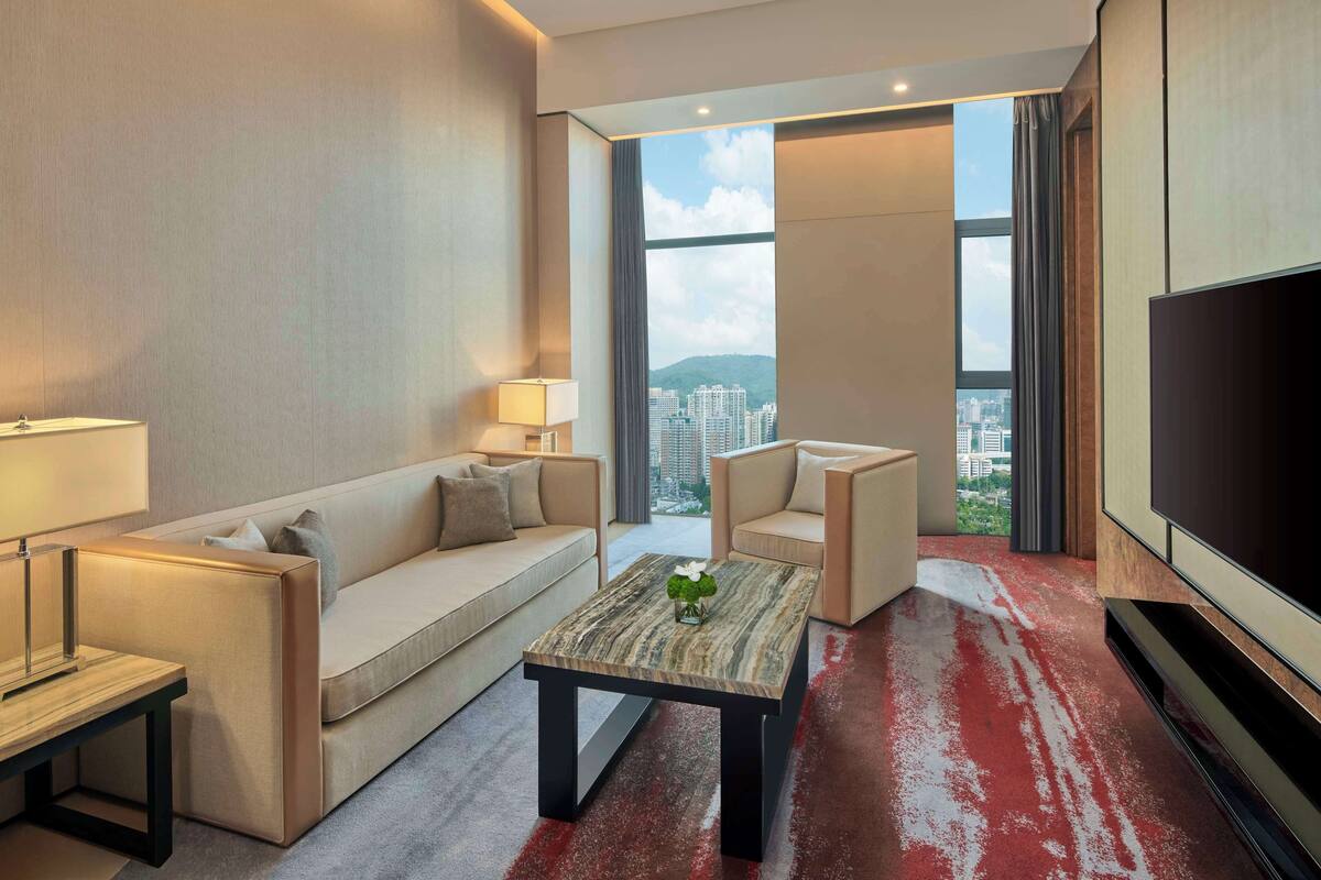 A modern hotel room with a sofa, a coffee table, a TV, and a large window offering a city view.
