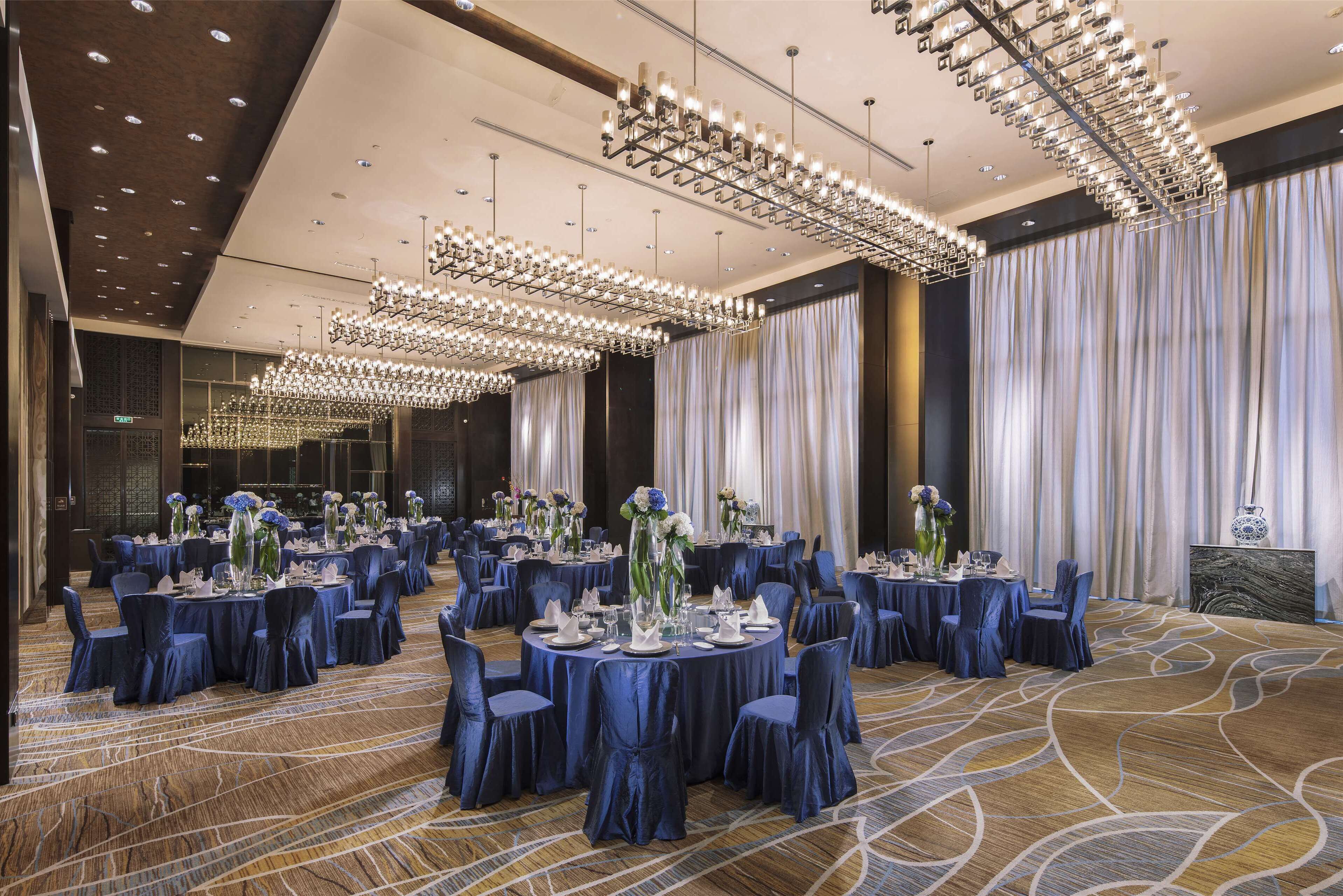 A banquet hall with round tables, blue chairs, and chandeliers.