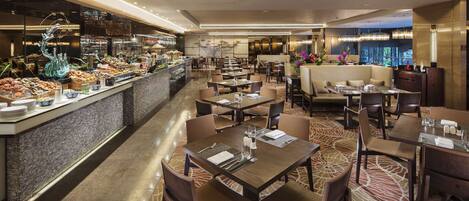 Un restaurante moderno con un bufé a la izquierda, varias mesas para comer con sillas y una fuente decorativa.
