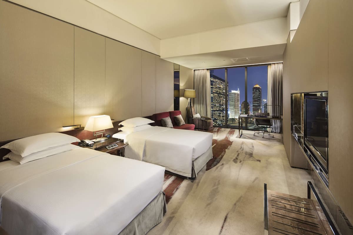 A hotel room with two beds, a TV, a desk with a lamp, and a city view.