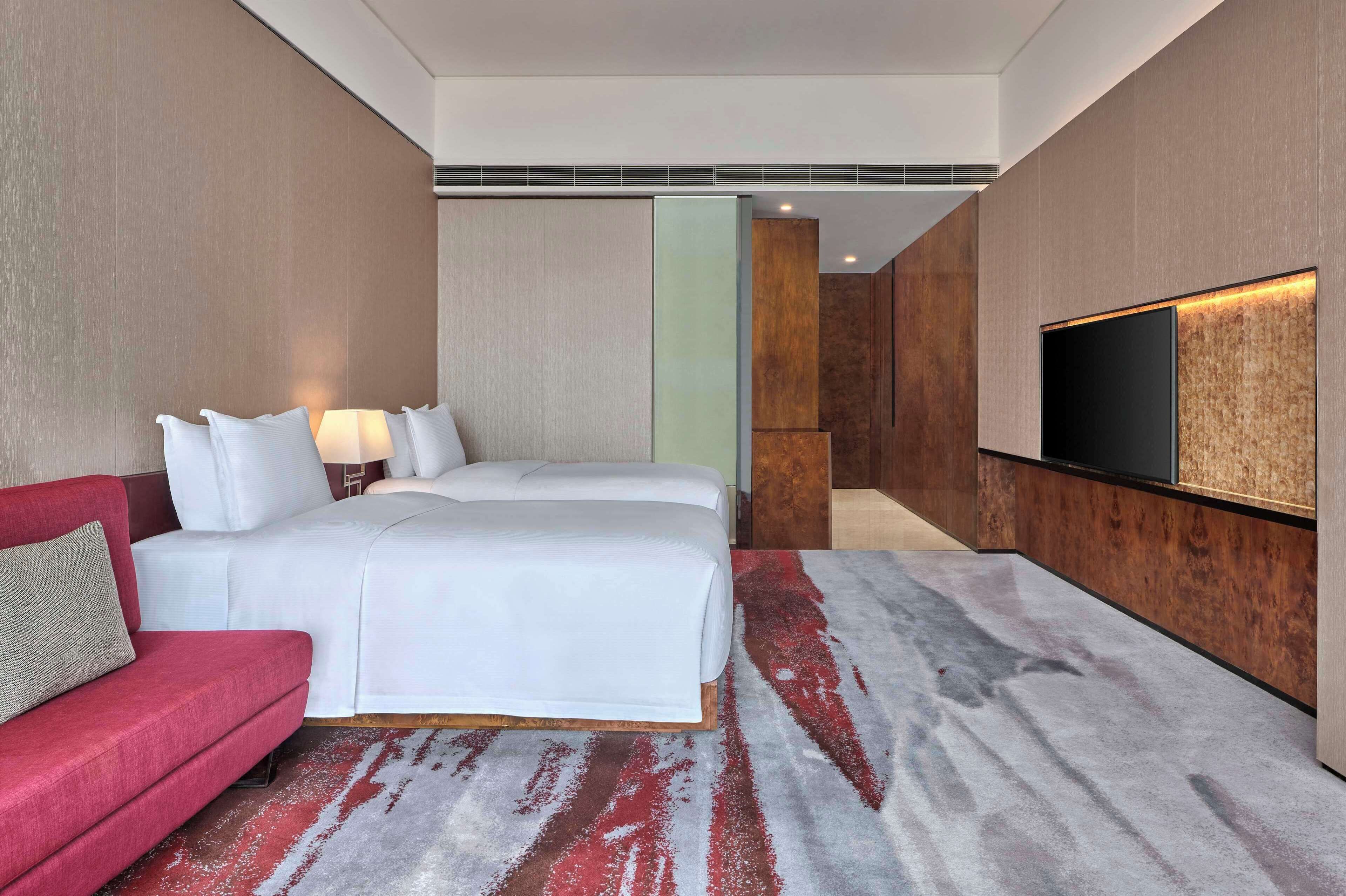 A hotel room with a large bed, a red sofa, a TV mounted on the wall, and a carpeted floor.