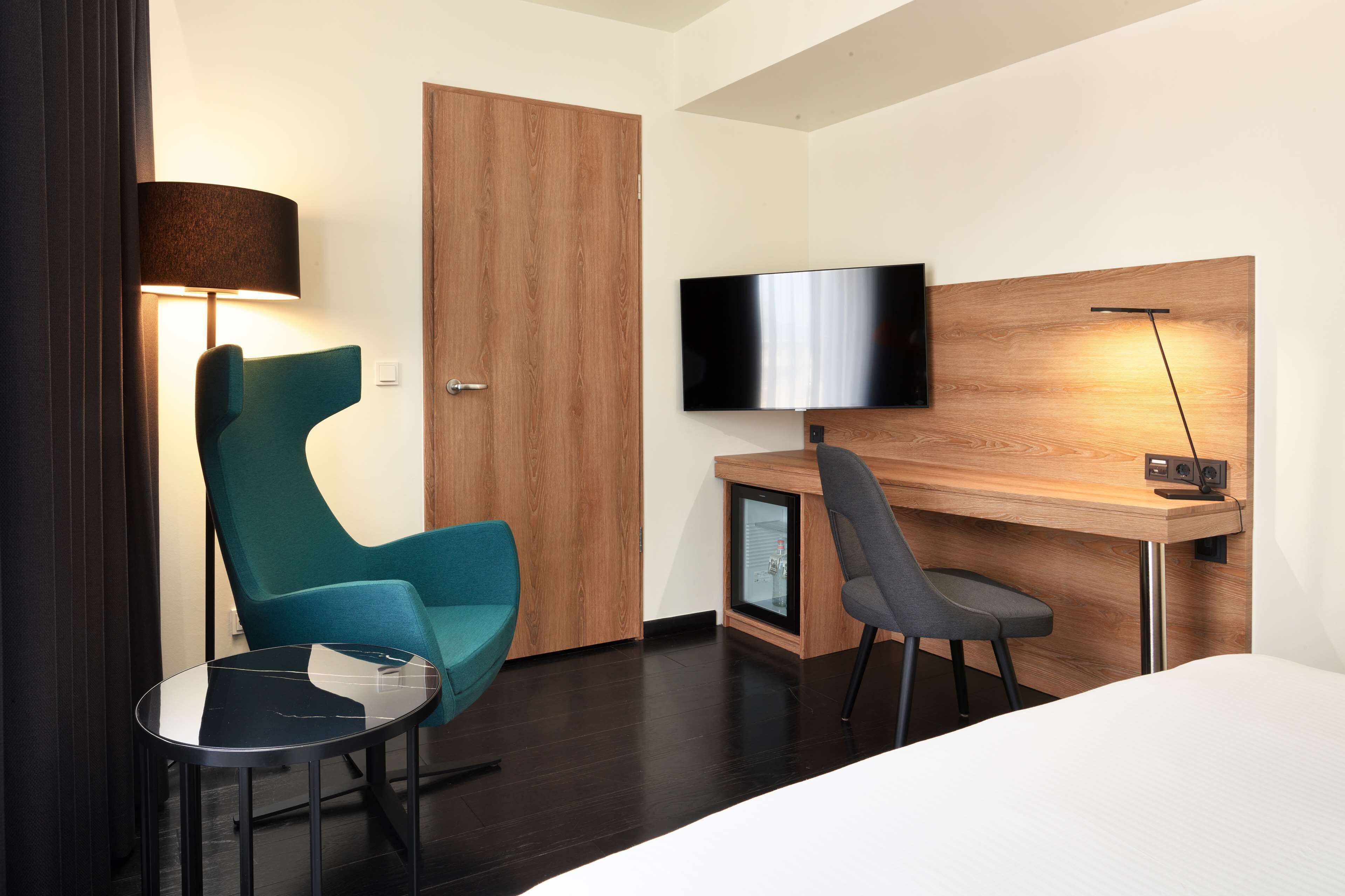 Habitación de hotel moderna con una cama, un escritorio con televisor, una silla verde azulada, una silla negra y una mesita.