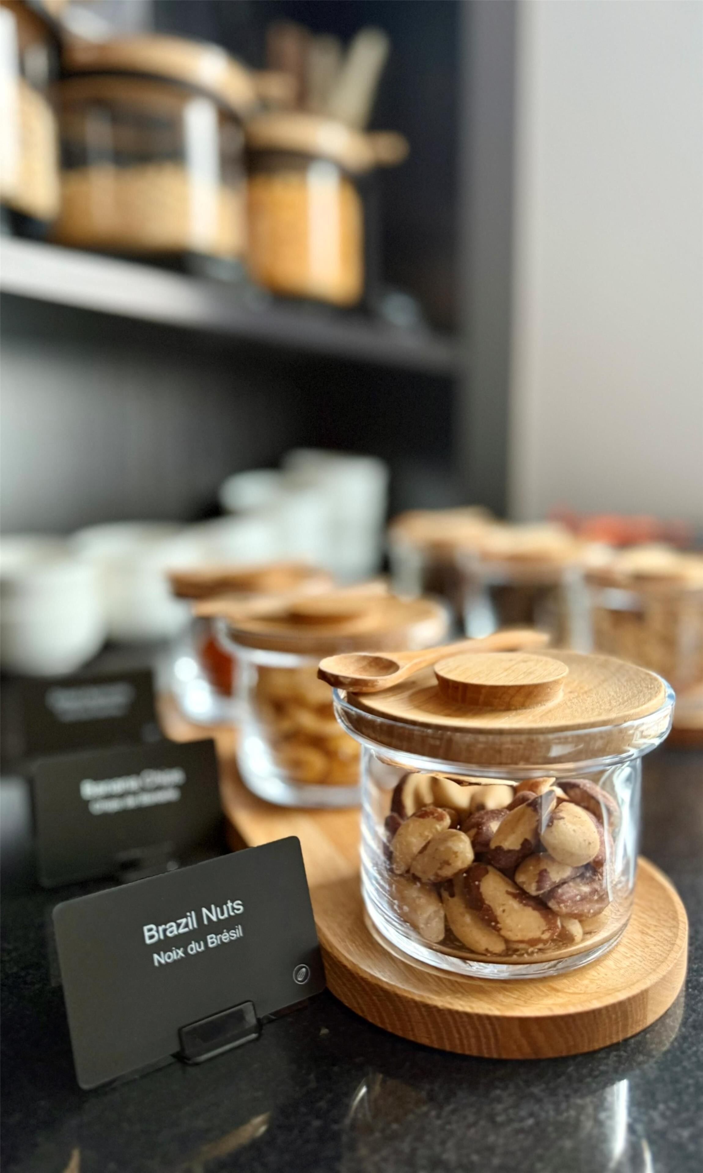A display of Brazil nuts in a glass jar with a wooden lid, labeled 'Brazil Nuts Noix du Brésil,' on a wooden board.