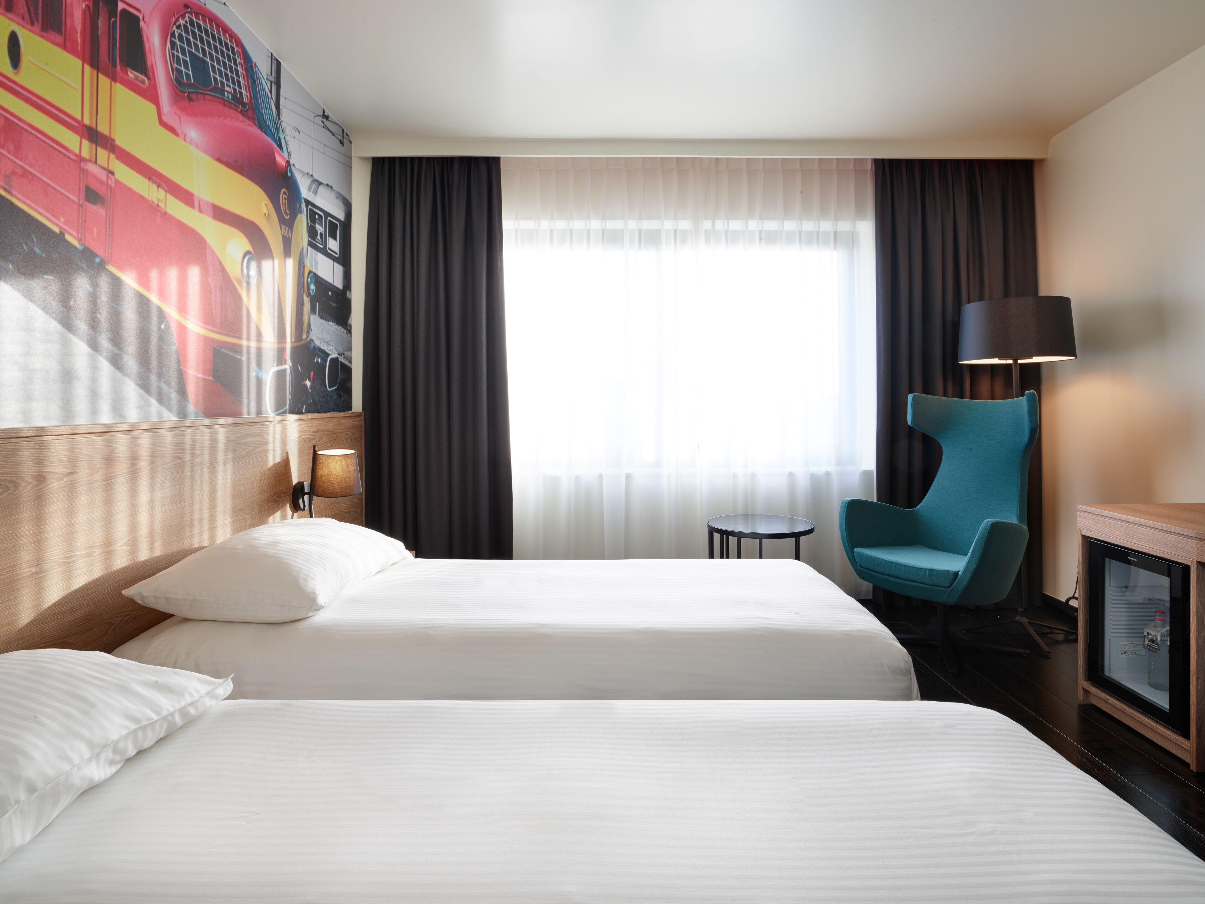 Habitación de hotel con dos camas, una silla, una mesita pequeña, una lámpara y un ventanal grande con cortinas.