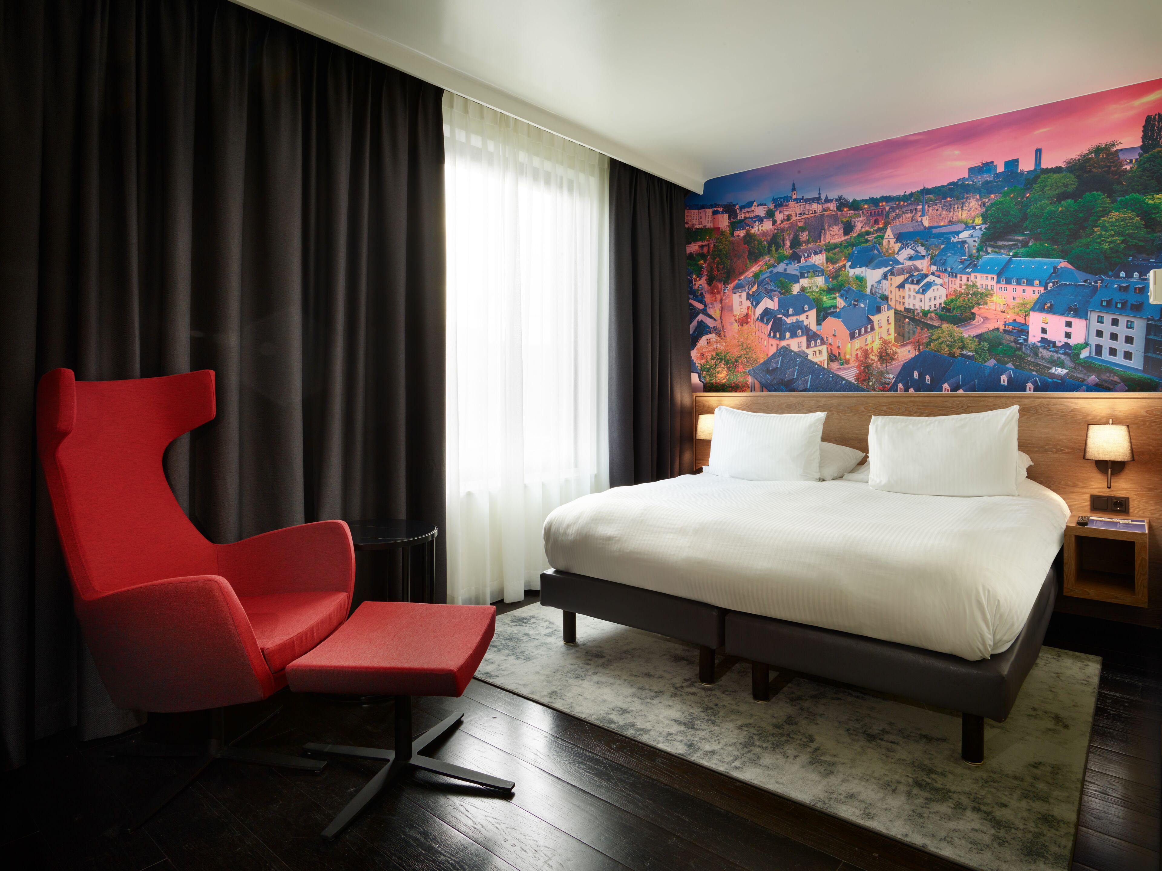 A hotel room with a large bed, a red chair, a cityscape mural on the wall, and a view of the city through the window.