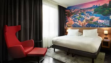 A hotel room with a large bed, a red chair, a cityscape mural on the wall, and a view of the city through the window.