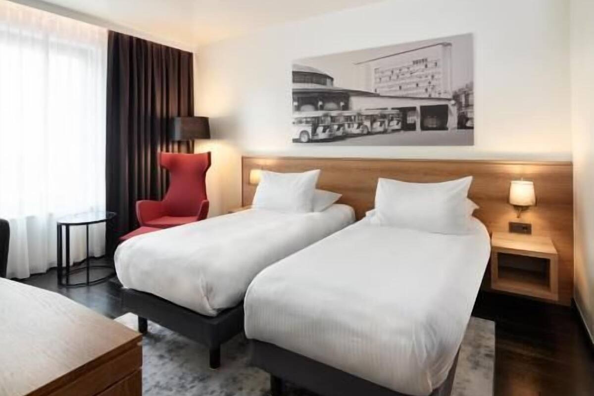 Una habitación de hotel con dos camas, una silla roja, una mesita y un cuadro colgado en la pared.
