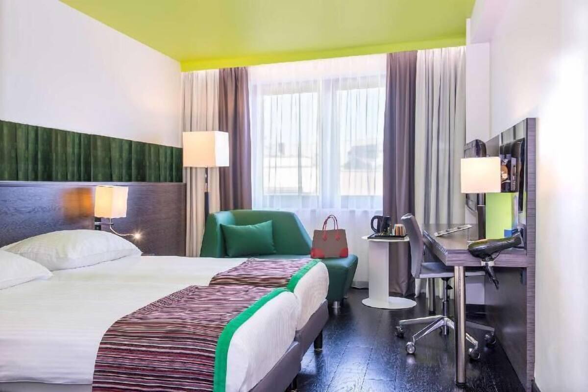 Habitación de hotel con cama, escritorio, silla, lámpara, cafetera y vista a la calle por la ventana.