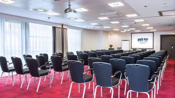 Un salón de conferencias con filas de sillas, una pantalla de proyector y un amplio ventanal con cortinas.