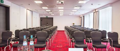 A conference room with rows of black chairs, a red carpet, a table with a phone, glasses, and water bottles.