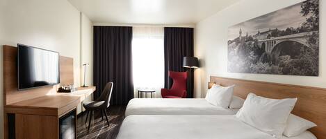A hotel room with two beds, a desk with a TV, a red chair, a lamp, and a large window with curtains.