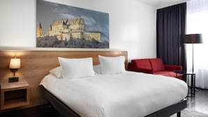 Habitación de hotel con una cama grande, un sillón rojo, una mesita de noche con una lámpara y un cuadro de un castillo en la pared.