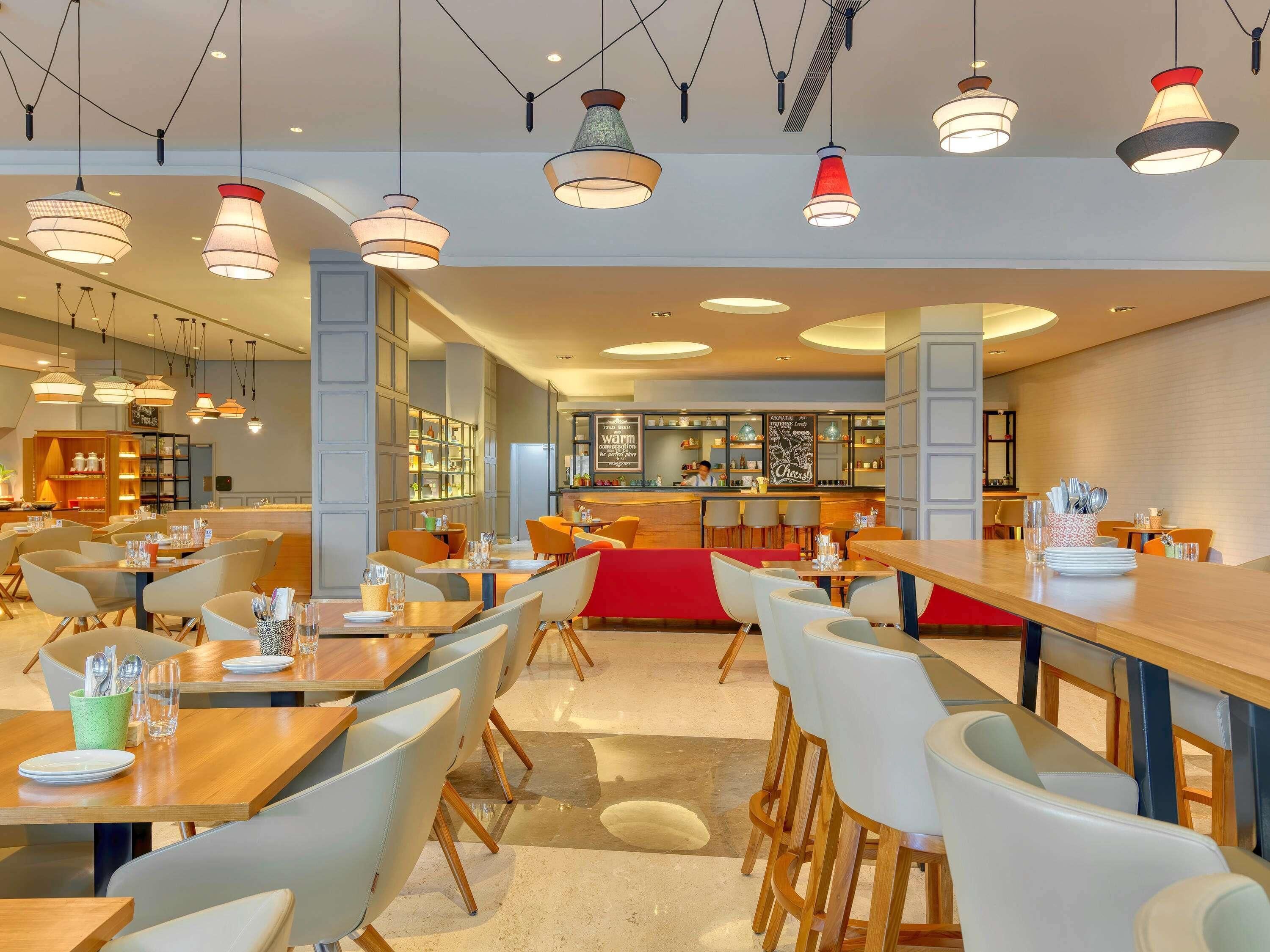 Et moderne restaurantmiljø med flere spiseborde og stole, hængende pendellamper og et barområde.