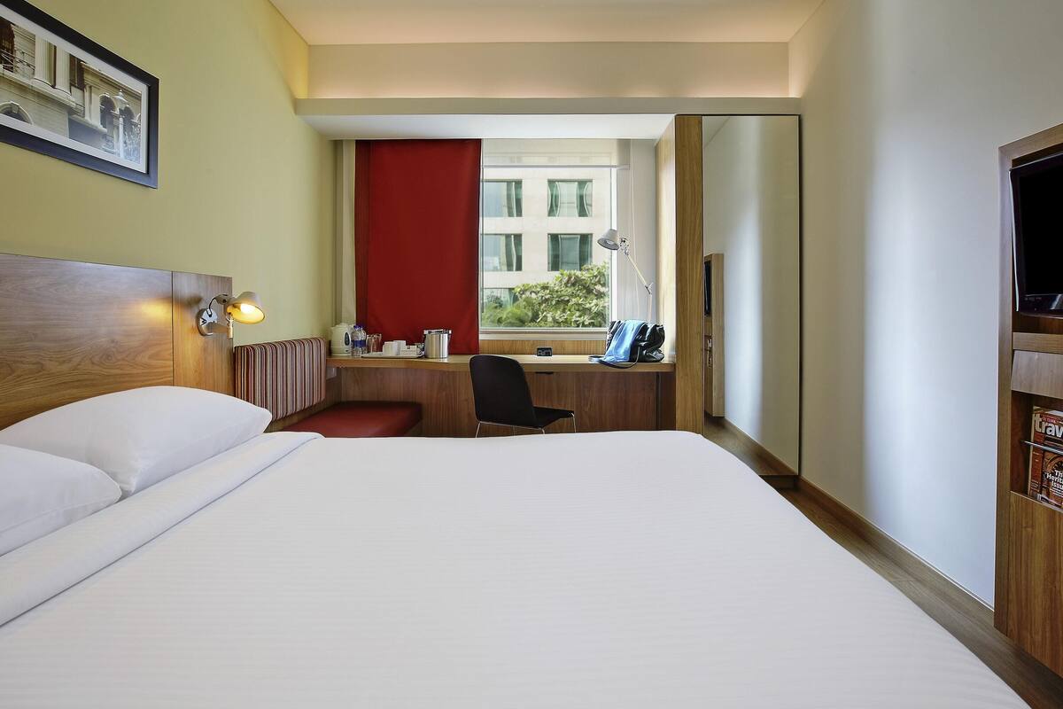 A hotel room with a bed, a desk with a chair, a window with a red curtain, a mirror, and a television.