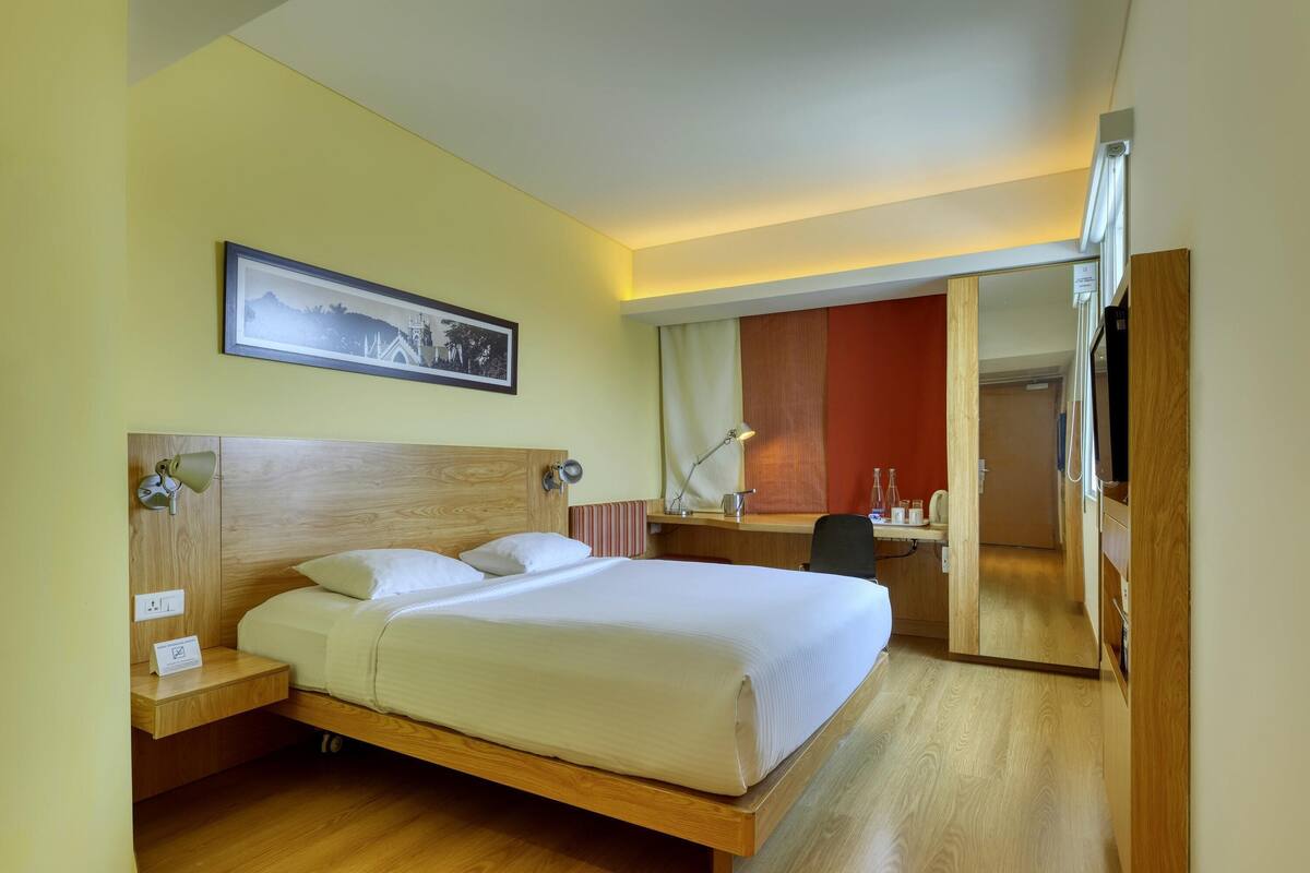 A hotel room with a wooden bed, a desk, a chair, a desk lamp, a framed picture on the wall, and a mirror.