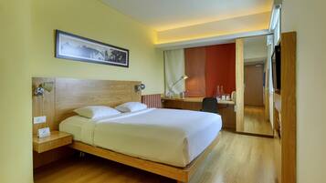 A hotel room with a wooden bed, a desk, a chair, a desk lamp, a framed picture on the wall, and a mirror.