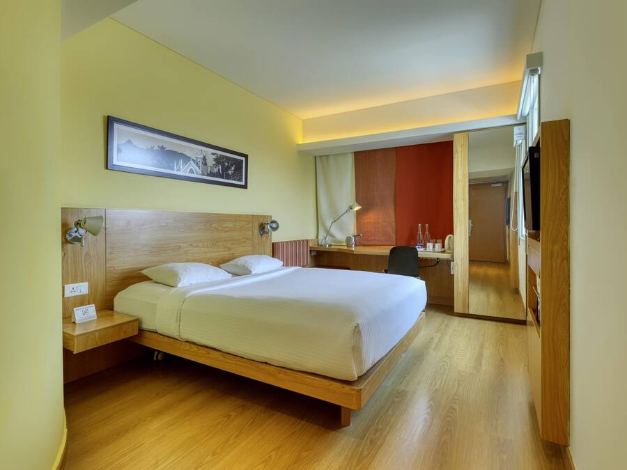 A hotel room with a wooden bed, a desk, a chair, a desk lamp, a framed picture on the wall, and a mirror.