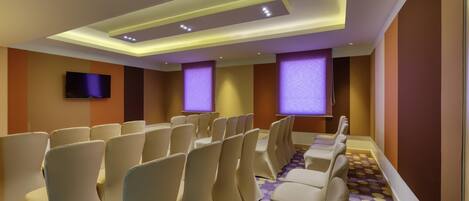 Une salle de conférence avec des rangées de chaises, un téléviseur à écran plat, des luminaires au plafond et une moquette à motifs.