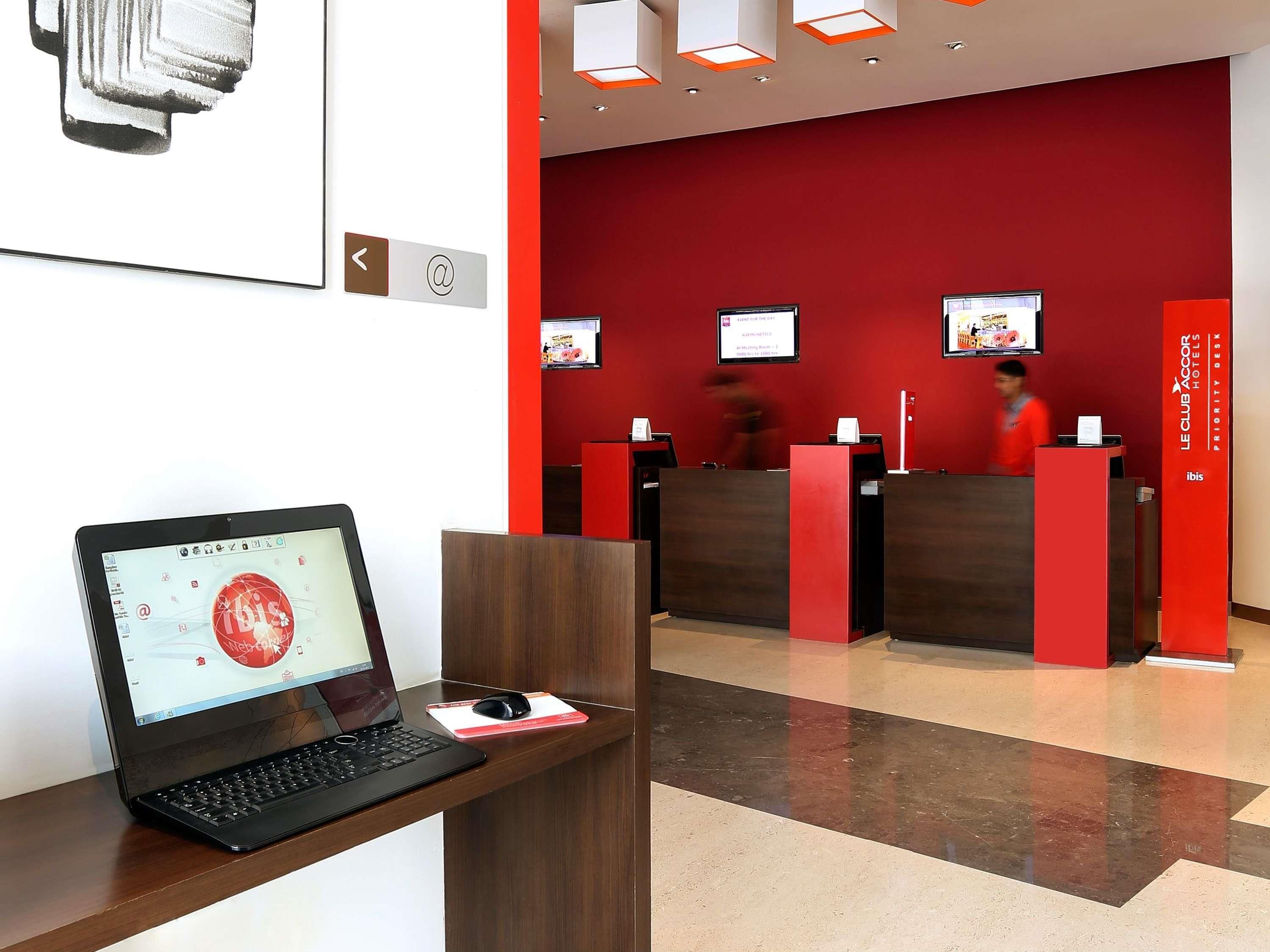 A hotel reception desk with a laptop and a mouse on a wooden stand, a red wall with a digital screen displaying a message, and a counter with a person in the background.