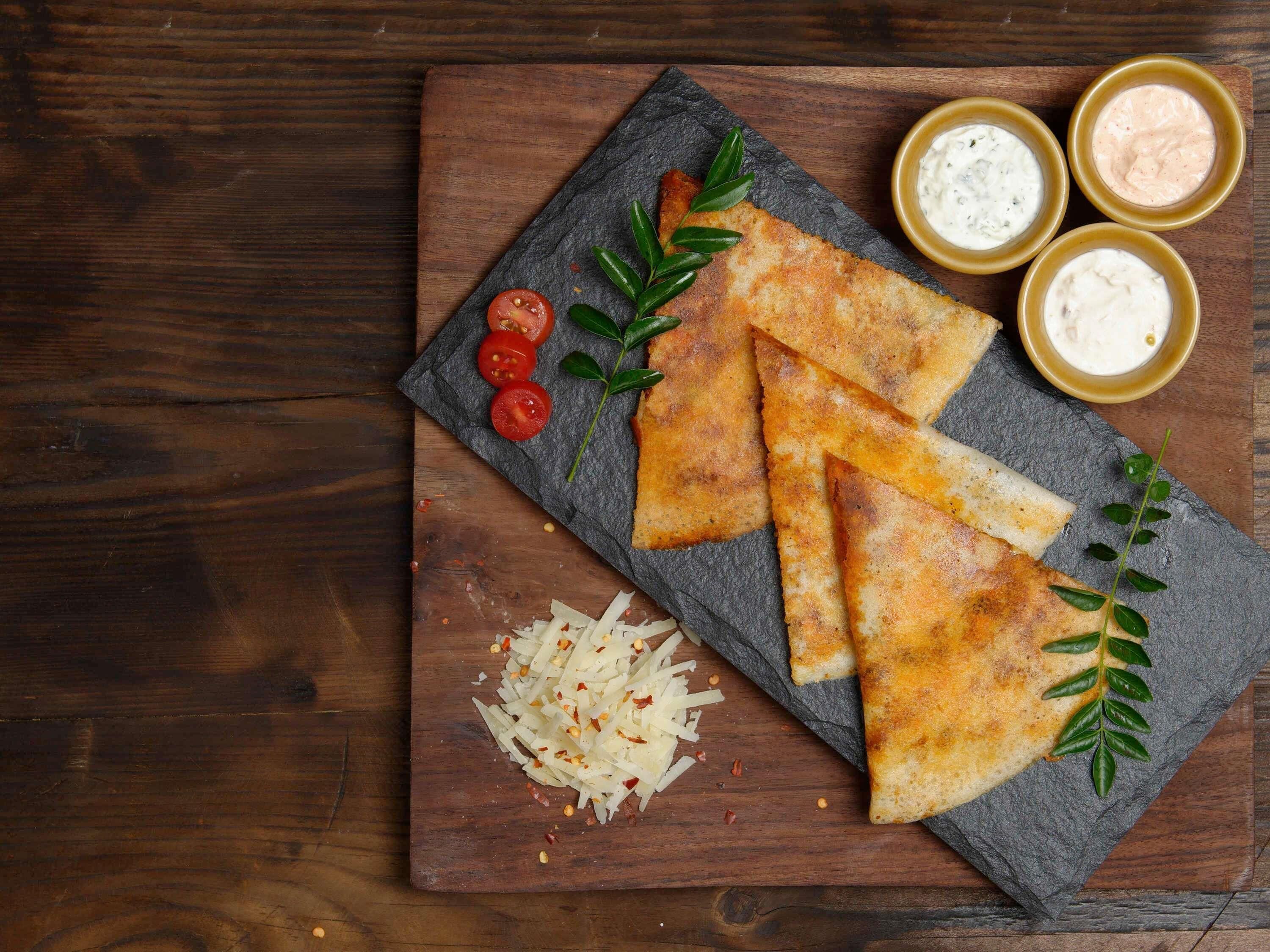 A slate board with triangular pieces of food, cherry tomatoes, shredded cheese, and two small bowls of sauce.