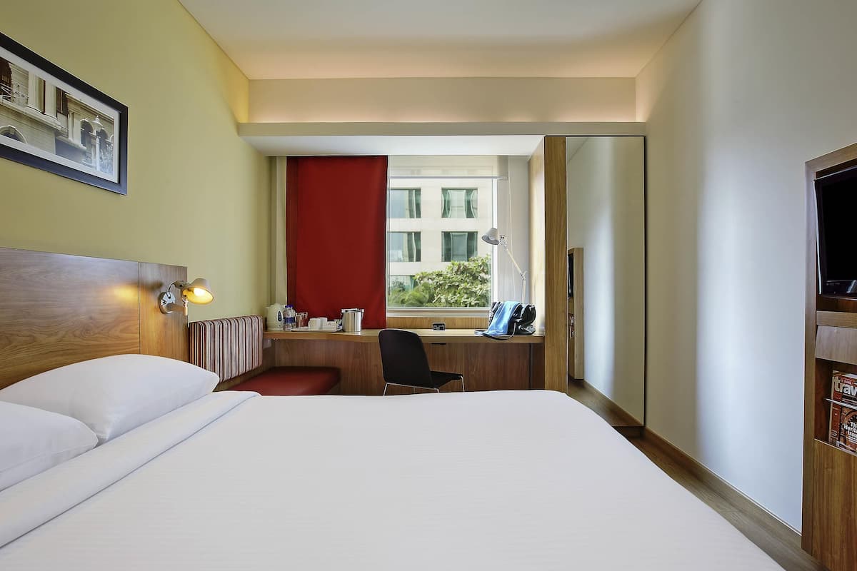 A hotel room with a bed, a desk with a chair, a window, and a mirror.