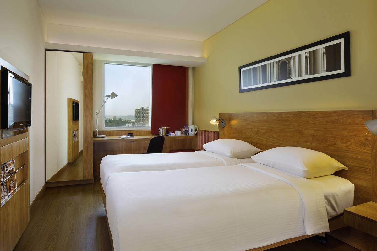 A hotel room with two beds, a desk, a TV, a window with a view, and a wooden headboard.