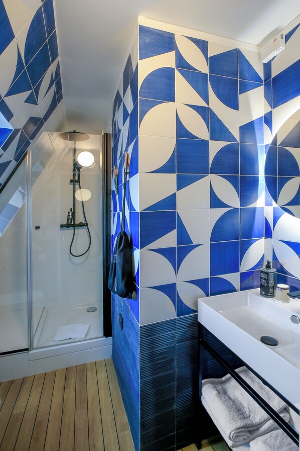 Un baño con azulejos azules y blancos, una cabina de ducha de vidrio, suelo de madera, un lavamanos blanco y un perchero negro.