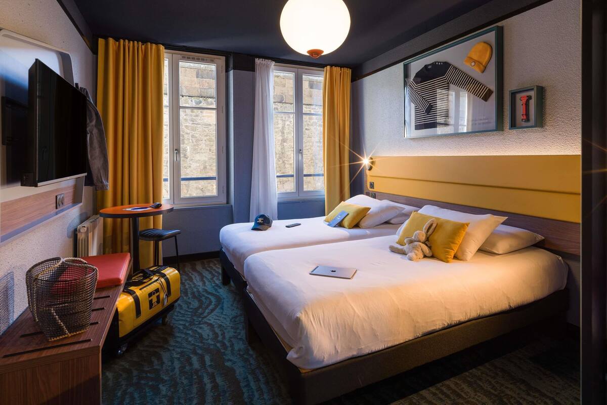 A hotel room with a bed, a desk, a chair, a suitcase, a TV, and a window with curtains.