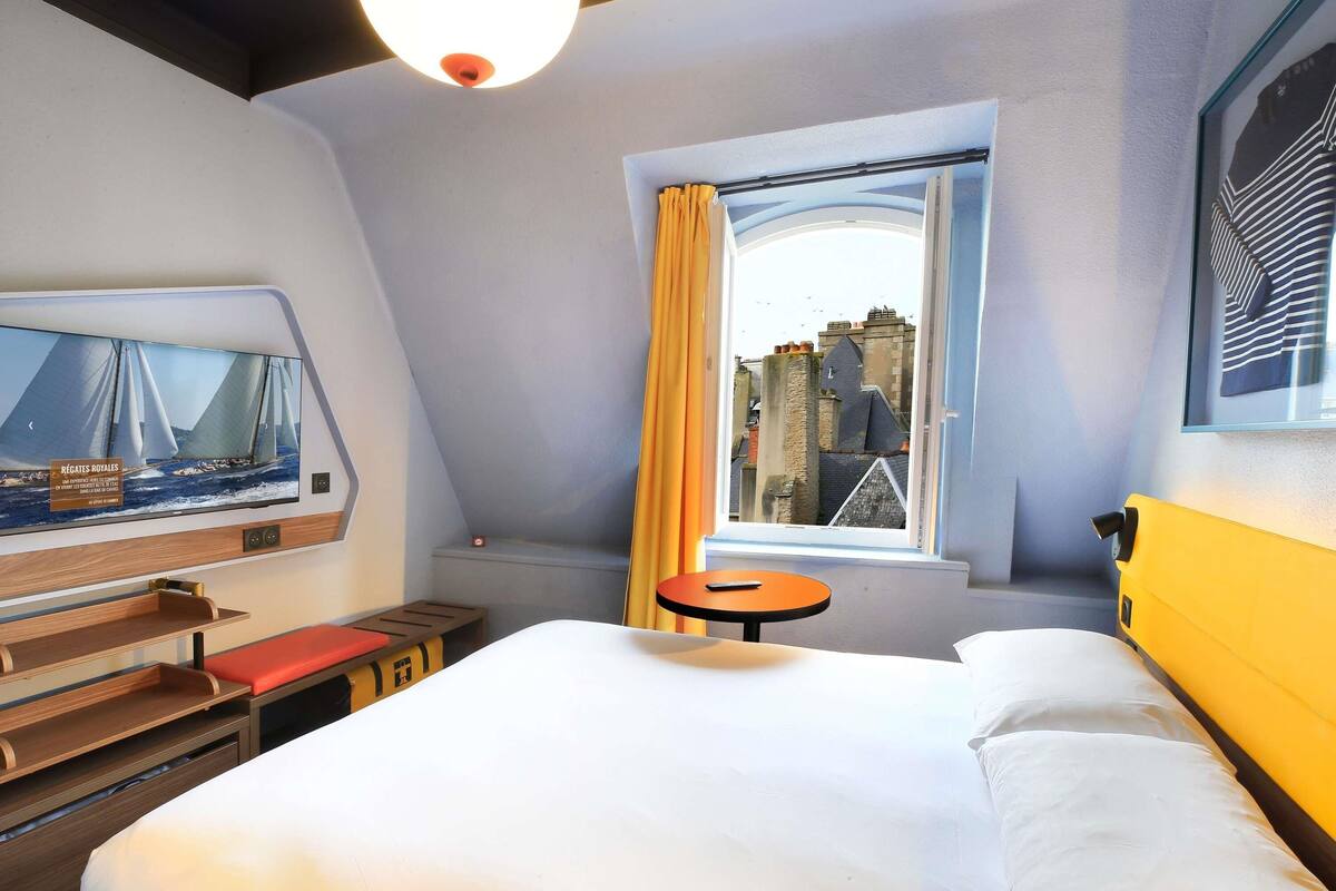 A hotel room with a bed, a small table, a TV mounted on the wall, and a window with a view of buildings.