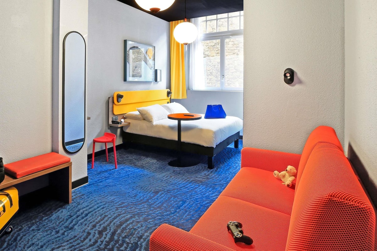 A hotel room with a bed, a red sofa, a small table, a chair, a mirror, and a window with curtains.