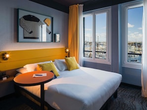 A hotel room with a bed, a small table, a nightstand, a framed picture on the wall, and a window with a view of a marina. - ibis Styles Saint-Malo Centre Historique (Saint-Malo)