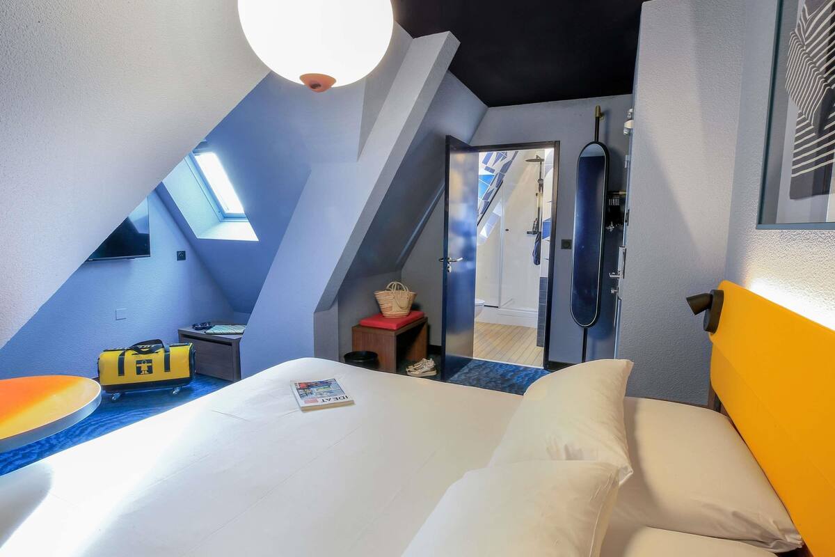 A bedroom with a bed, a bedside table, a lamp, a window, and a door leading to a bathroom.