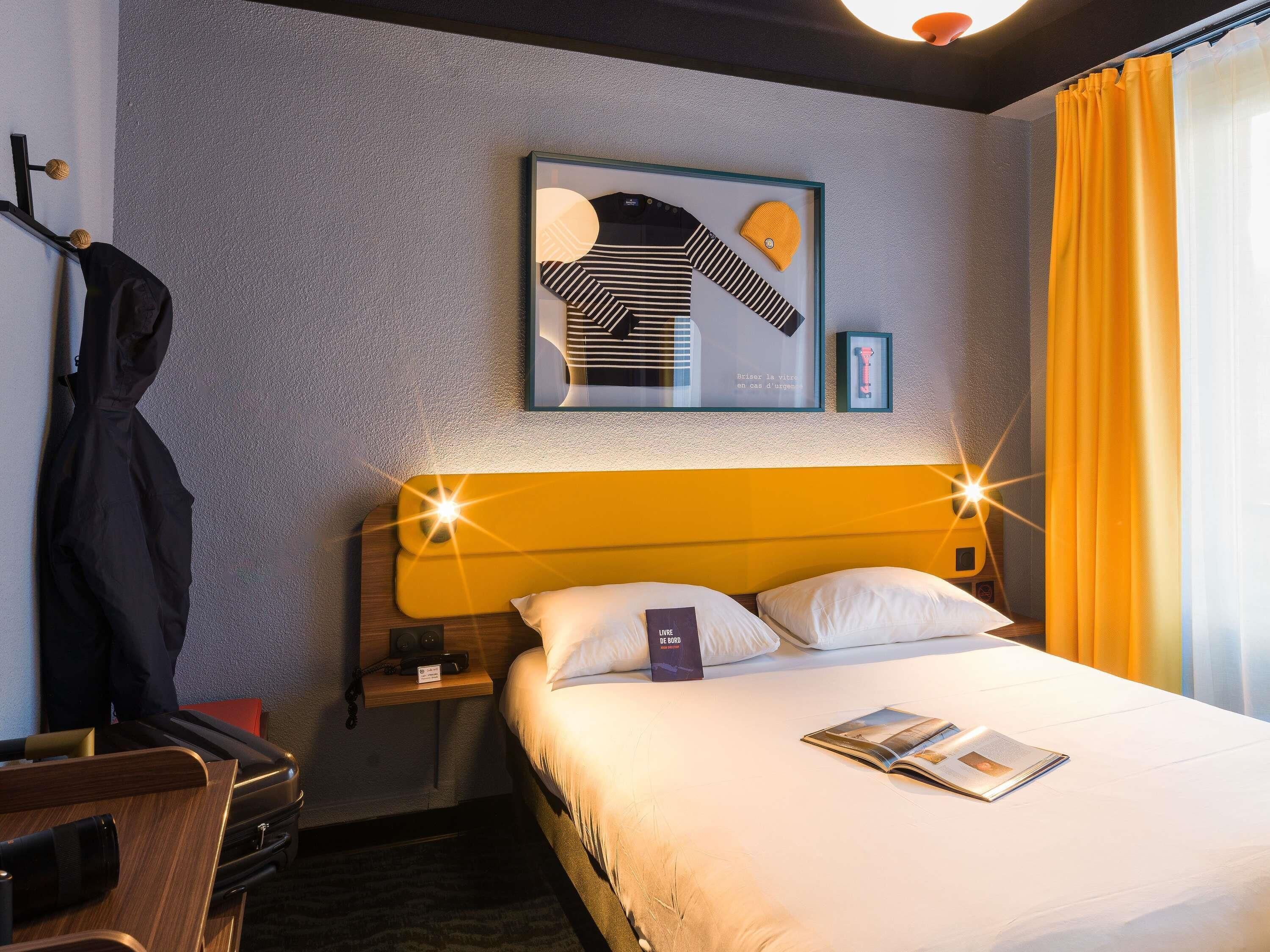 Quarto de hotel com cama, mesa de cabeceira, mala e uma obra de arte emoldurada na parede.