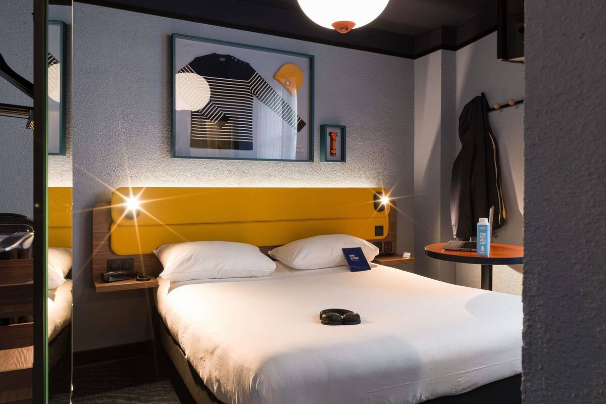 A hotel room with a bed, a bedside table, a lamp, a framed picture on the wall, and a coat hanging on the wall.