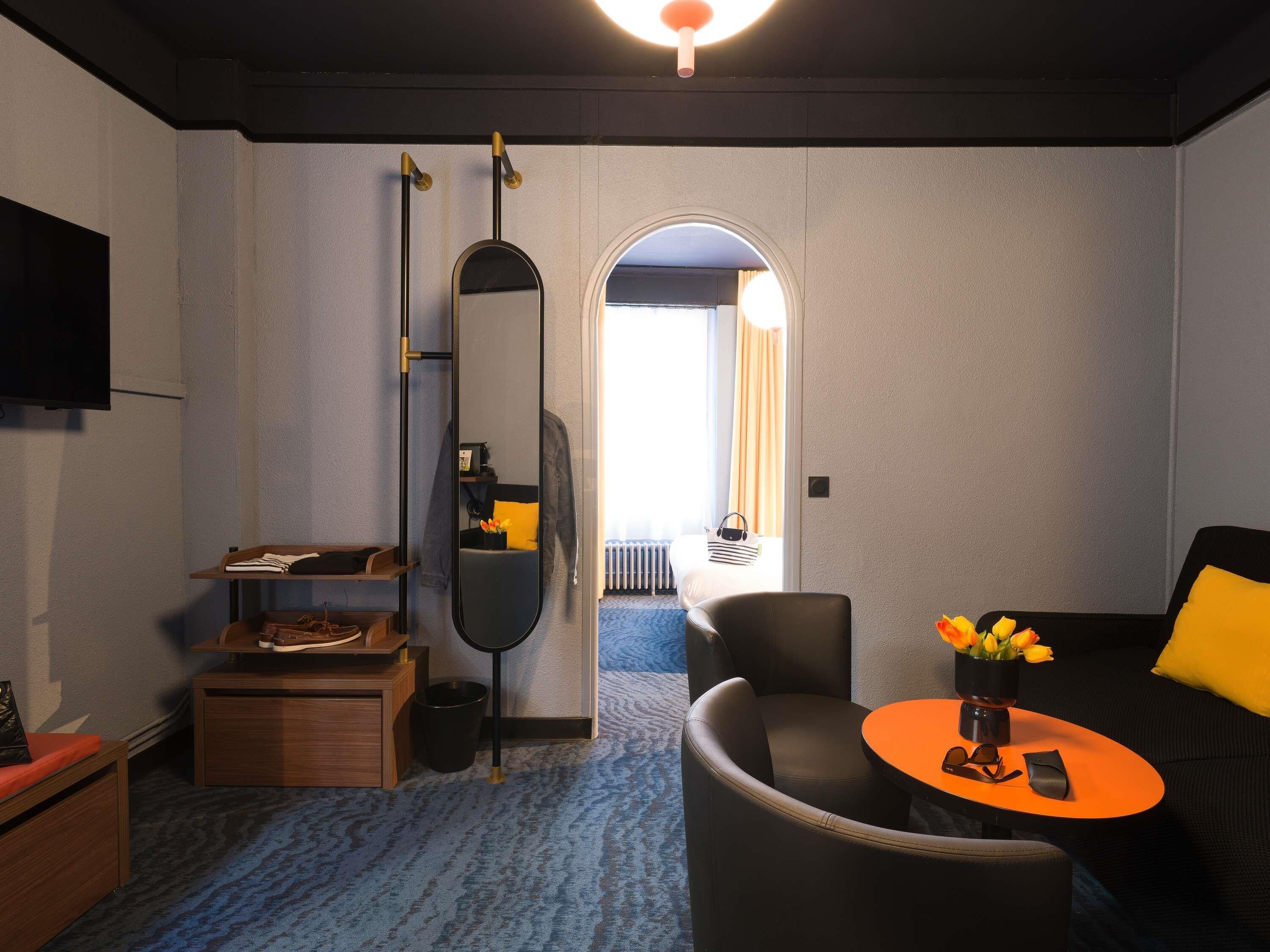 Una moderna camera d'albergo con un ampio specchio, un tavolino rotondo con un vaso di fiori, un divano con un cuscino giallo, un comodino in legno e un televisore a parete.