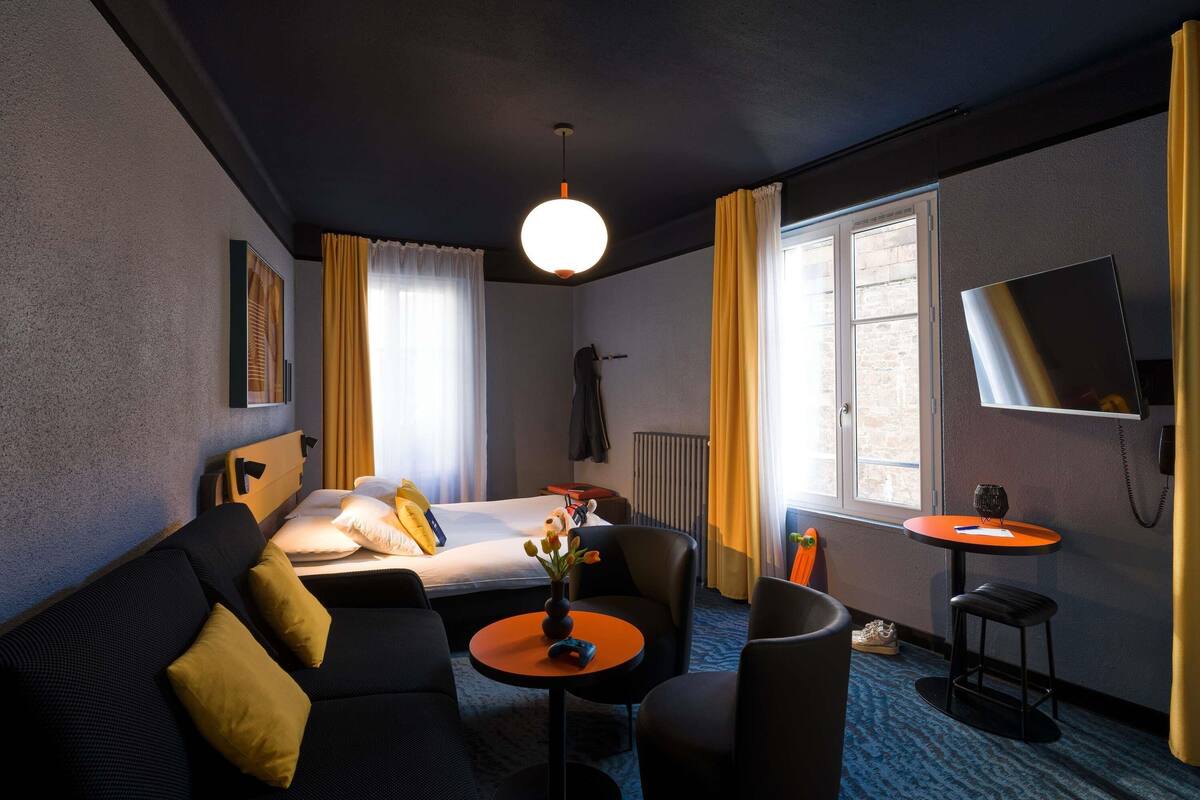 A hotel room with a bed, sofa, armchair, small round table, TV, and a window with curtains.