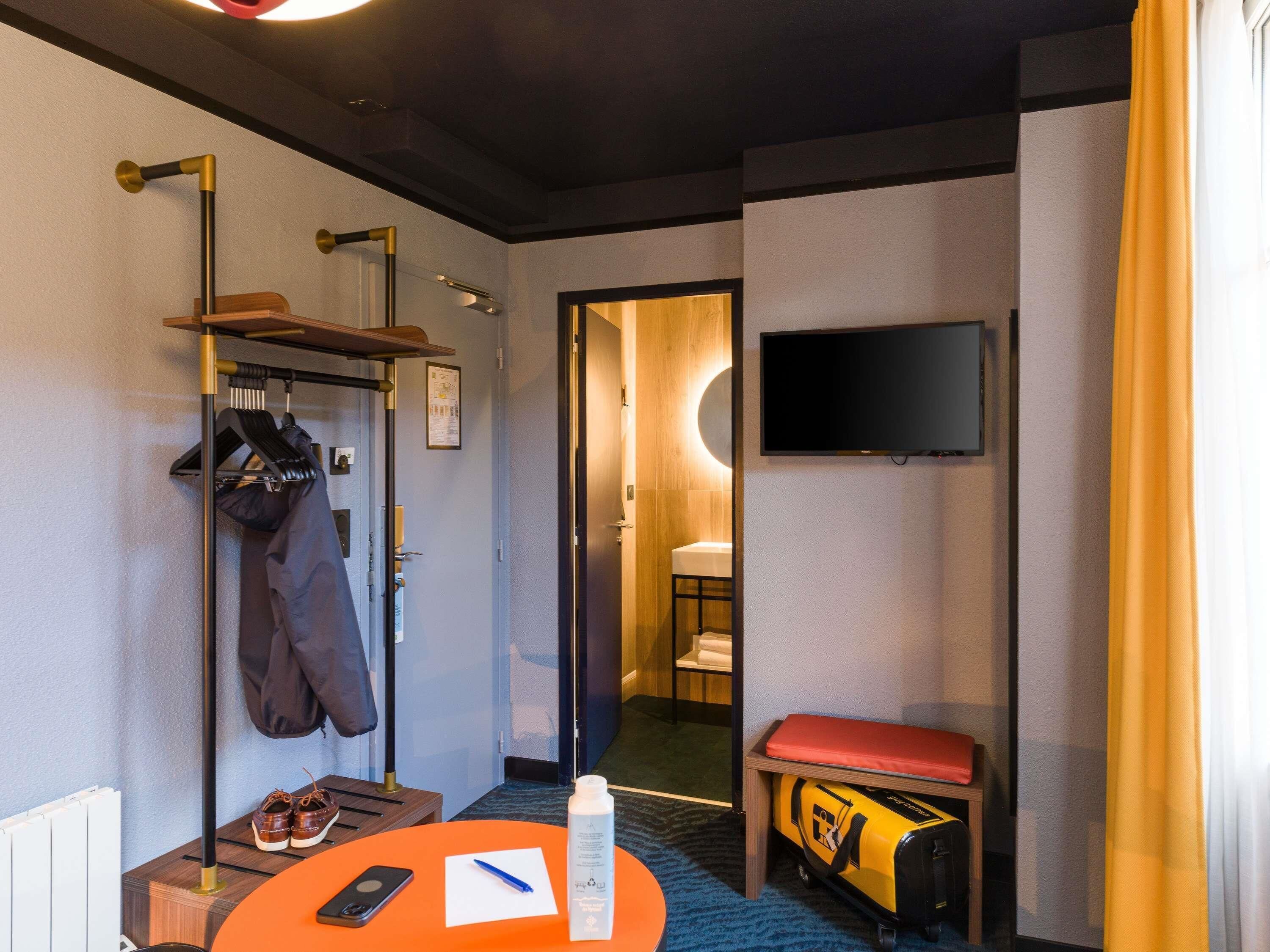 Habitació d'hotel amb una perxa, televisió, una panxa, taula amb un telèfon i una ploma, i un bany visible a través d'una porta oberta.