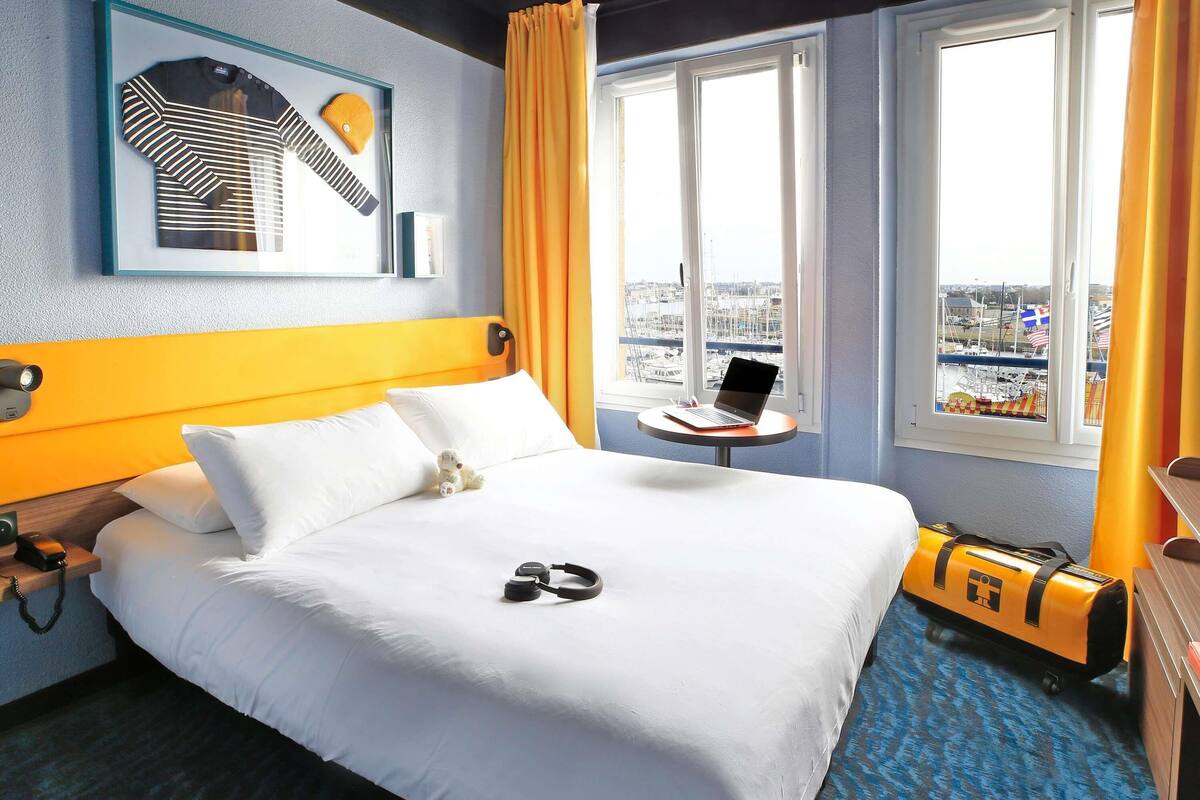 A hotel room with a bed, a small table with a laptop, a teddy bear, and a pair of headphones on the bed.