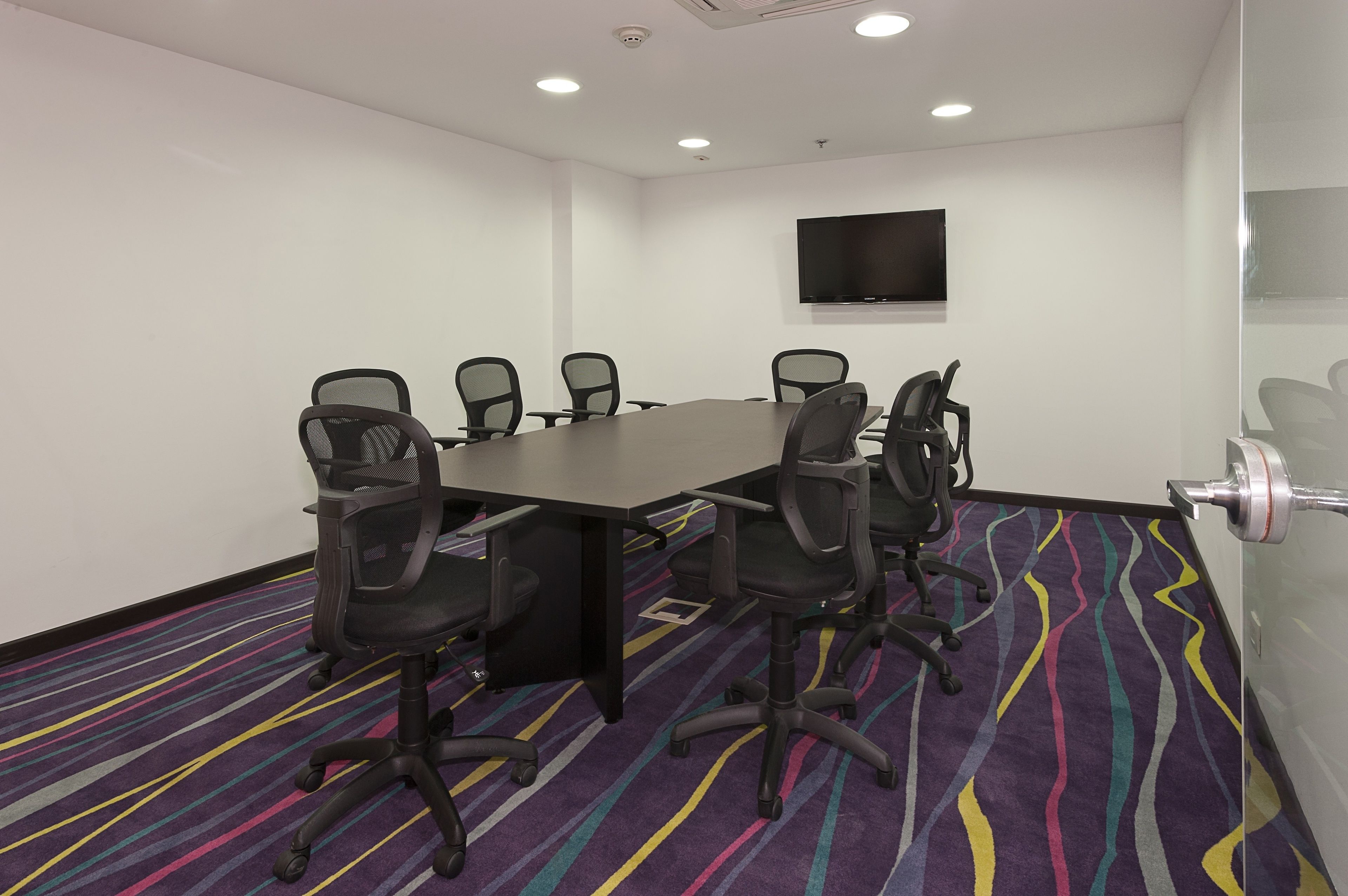 Une salle de conférence avec une longue table, six chaises, un téléviseur à écran plat fixé au mur et un tapis rayé aux couleurs vives.