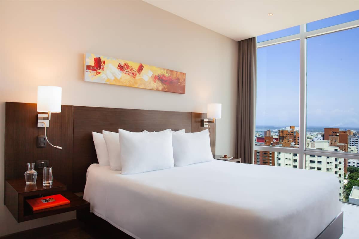 A hotel room with a large bed, two bedside lamps, a nightstand with a red book, a painting on the wall, and a city view through large windows.